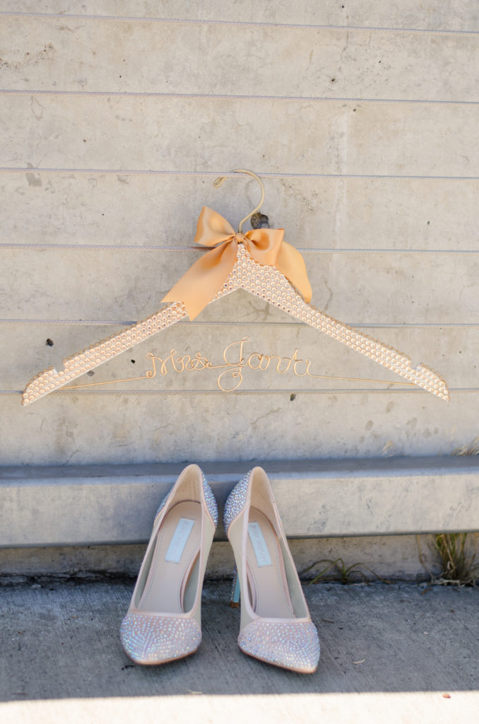 Wooden hanger embellished with sparkled gems and caramel ribbon in a bow with sparkled heels below and concrete as a backdrop 